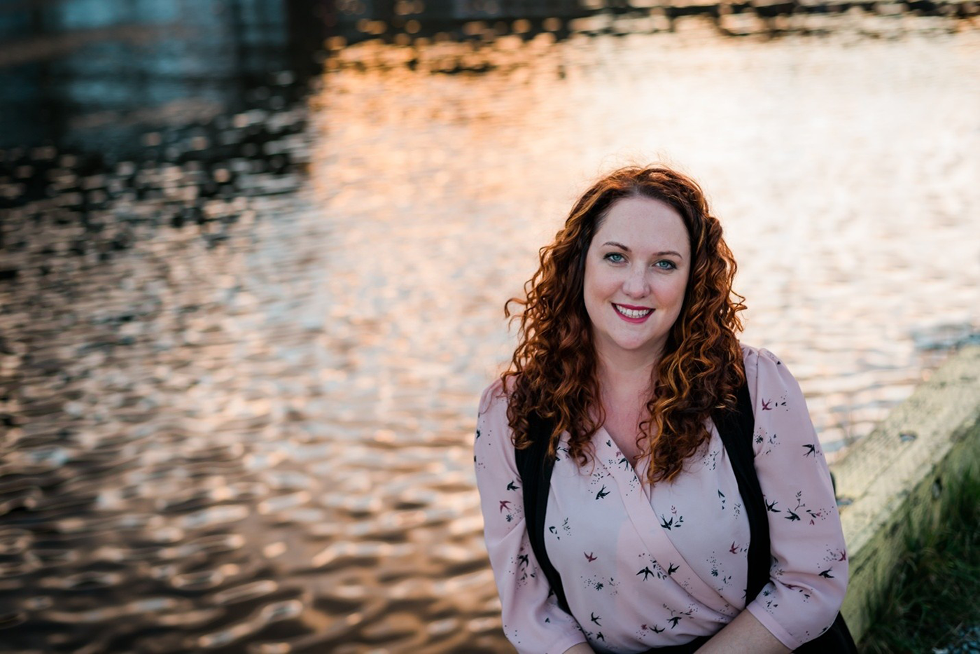 A woman with curly, red hair wearing a pale pink shirt smiles at the camera and sits in front of a body of water at sunset.