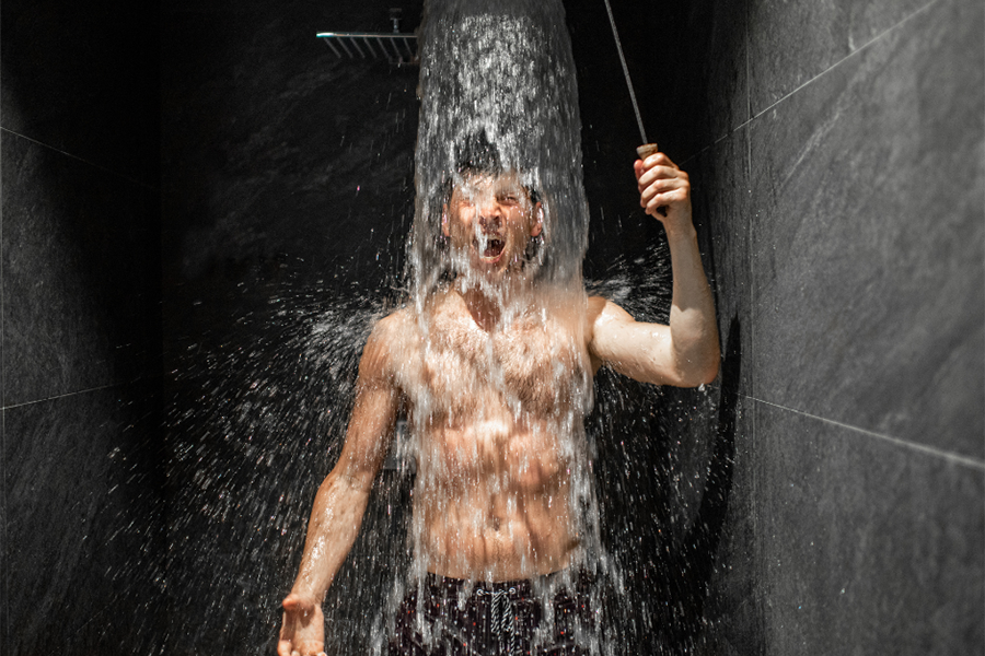 A man stands shirtless under a heavy stream of water coming from the ceiling inside a grey shower.