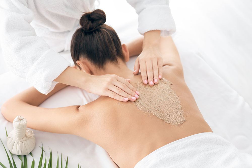 A dark-haired woman lies face down on a massage table while a person rubs scrubbing salts on her bare back.