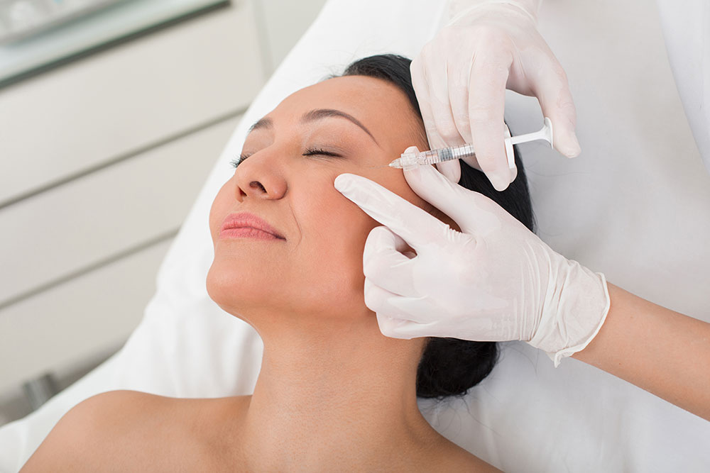 A dark-haired woman lies on her back with her eyes closed while a person wearing white medical gloves injects a syringe into her forehead.