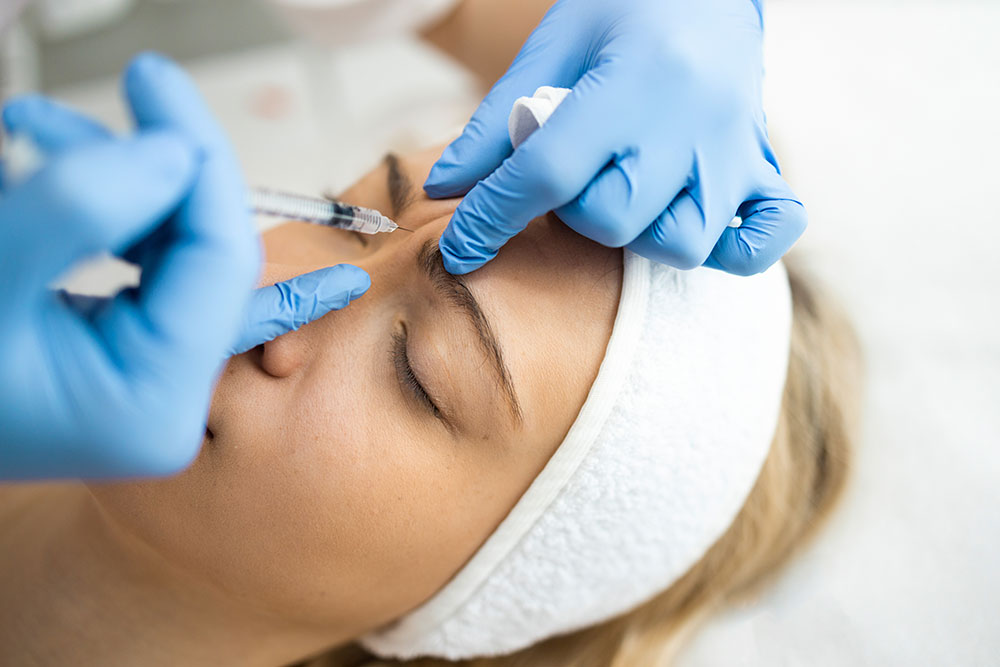 A blond woman wearing a white headband lies on her back with her eyes closed while a person wearing blue medical gloves injects a syringe into her forehead.