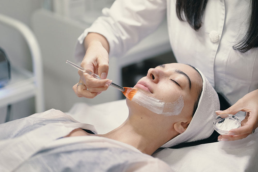 A dark-haired woman wearing a white headband lies on her back with her eyes closed while a person paints a white substance on her face with a paintbrush.