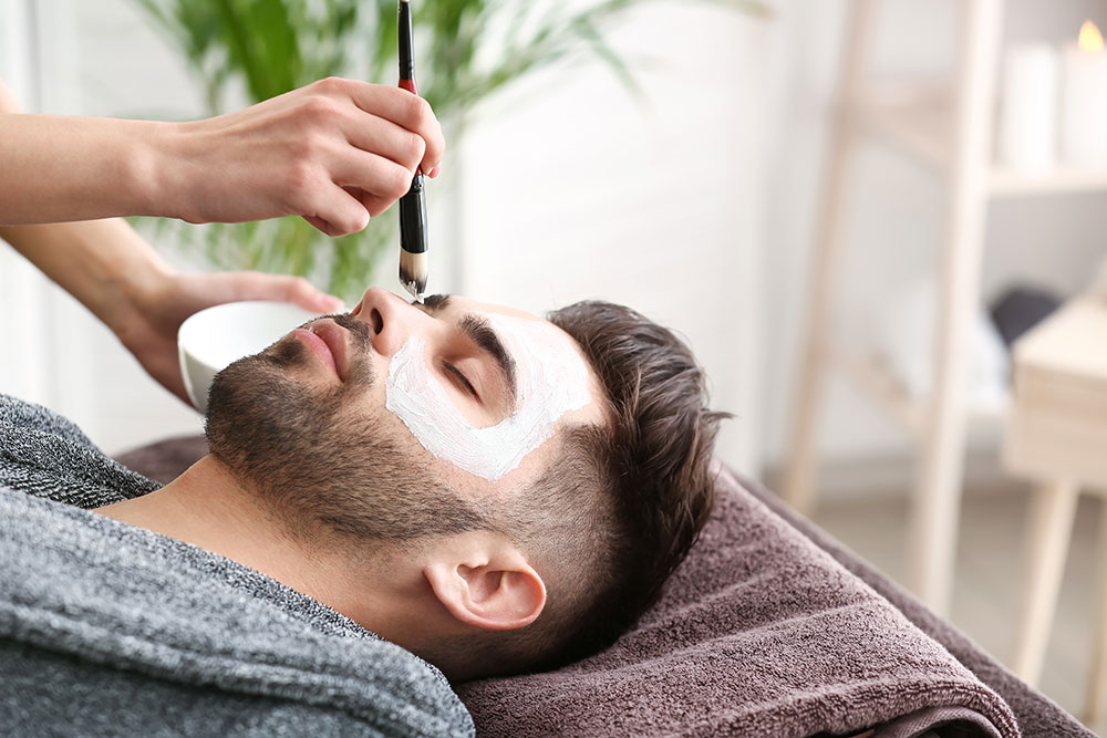 A brown-haired man with a trimmed beard lies on his back with his eyes closed while a person paints a white substance on his face with a paintbrush.