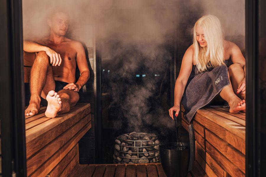 A man and a woman sit inside a wooden sauna with steaming stones in between them.