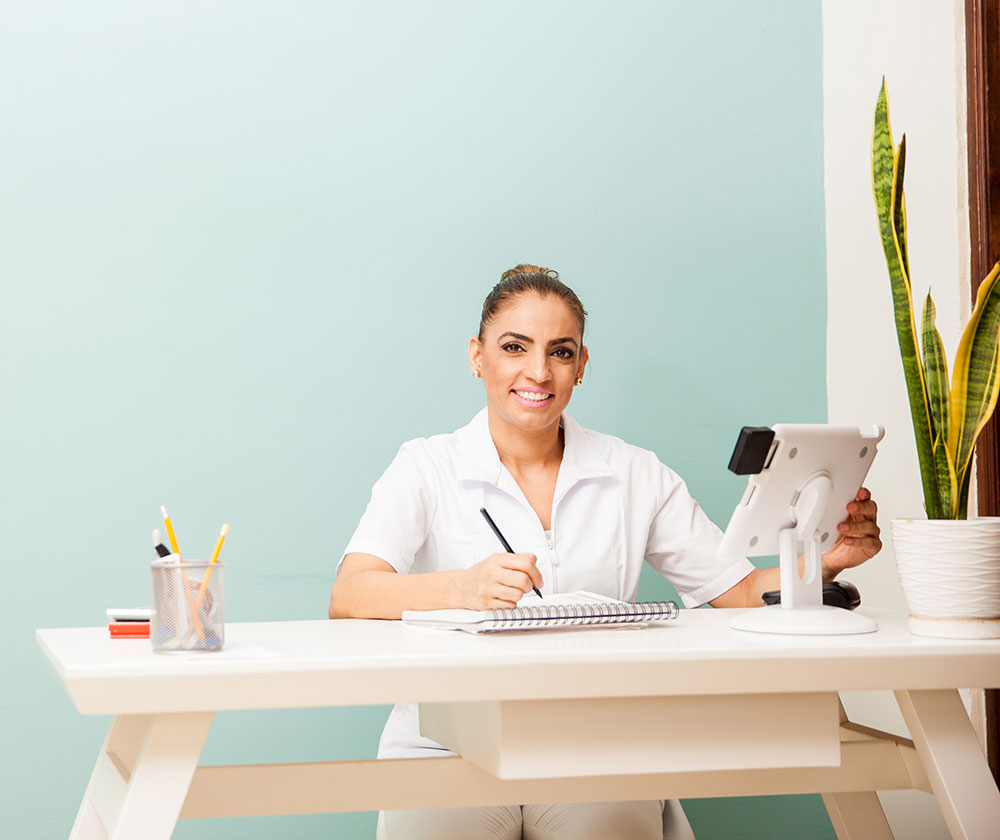 A woman in a white, short-sleeved uniform sits at a desk with a notebook, a digital tablet, a container of pencils and a potted plant in a pale blue painted room.